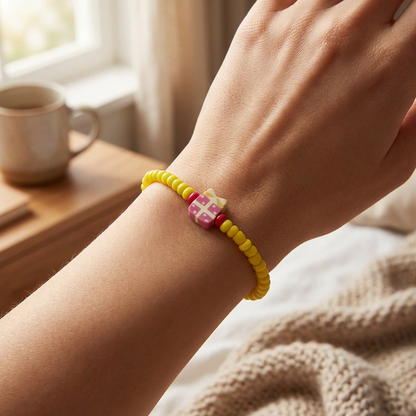 Hand wearing a yellow beaded bracelet with a pink and red heart charm, in a cozy indoor setting.