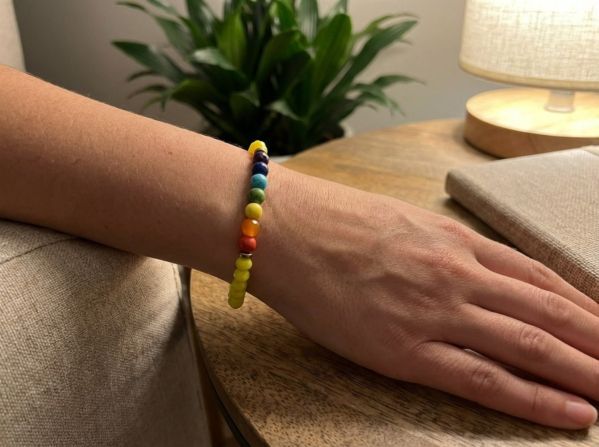 Hand wearing a colorful beaded bracelet on a wooden surface with a plant and lamp in the background