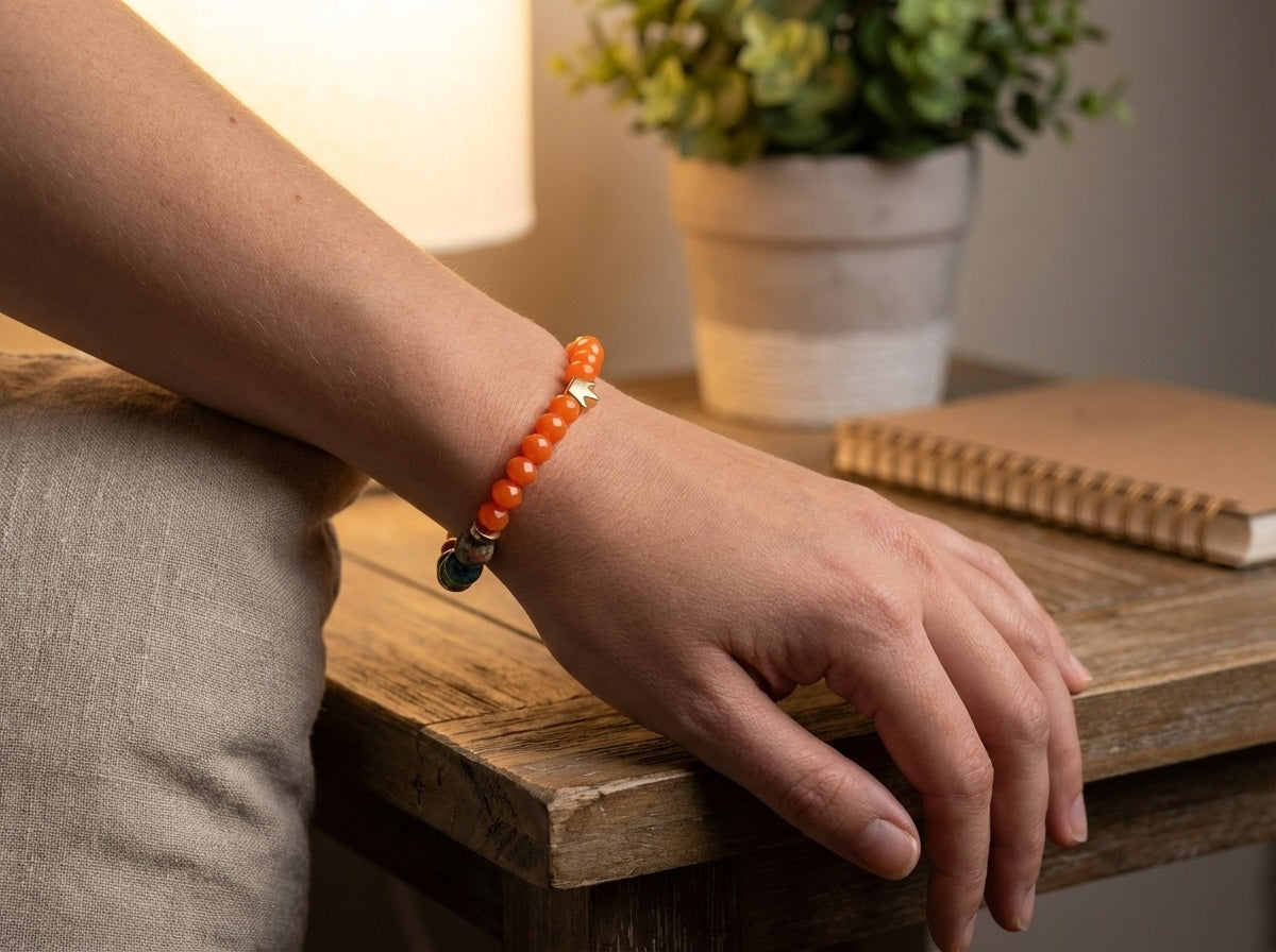 Person wearing an orange beaded bracelet on a wooden surface with a plant and notebook in the background.
