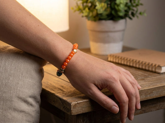 Person wearing an orange beaded bracelet on a wooden surface with a plant and notebook in the background.