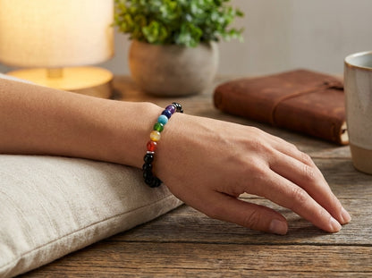 Hand wearing a colorful bracelet on a wooden surface with a lamp and plant in the background