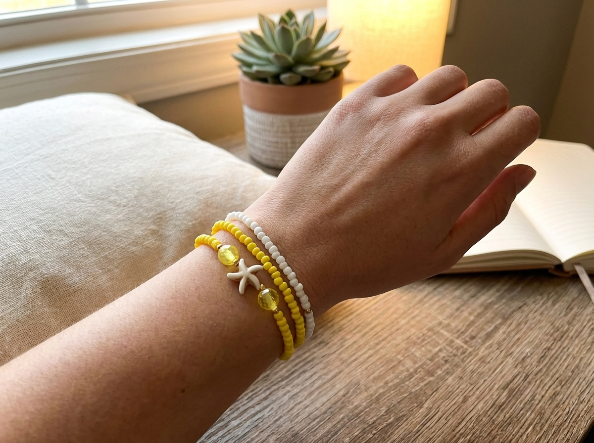 Hand wearing a yellow and white beaded bracelet on a wooden surface with a book and plant in the background.