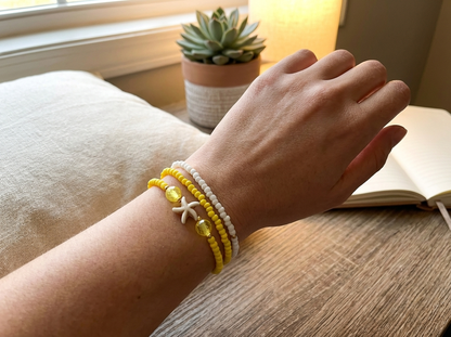 Hand wearing a yellow and white beaded bracelet on a wooden surface with a book and plant in the background.