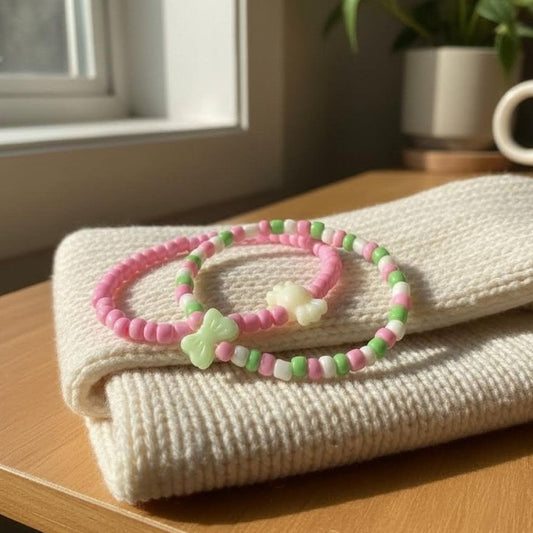 Two pink and green beaded bracelets on a folded beige blanket with a window and plant in the background.