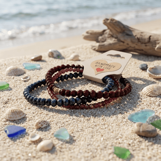 Three beaded bracelets on sand with a 'Handmade with Love' card, surrounded by shells and driftwood.