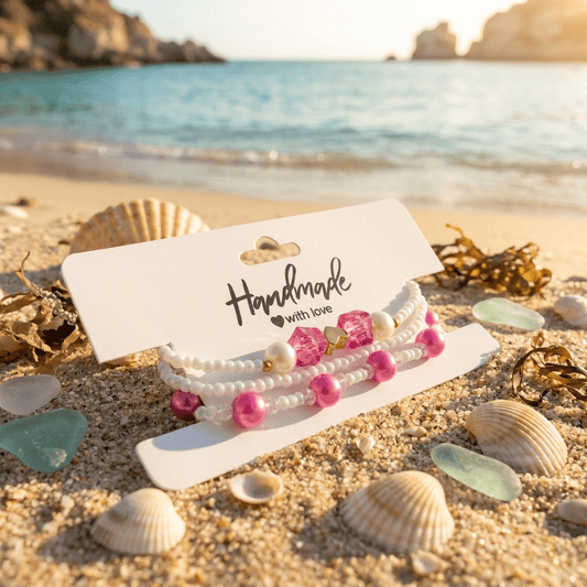 Set of pearl and pink beaded bracelets on a card with 'Handmade with love' text, placed on sand with seashells and ocean view.