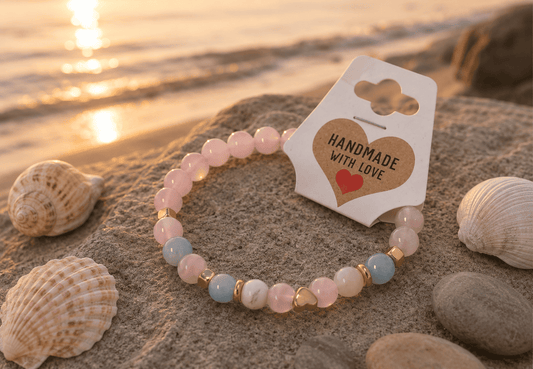 Beaded bracelet with 'Handmade with Love' tag on a beach with shells and sunset.