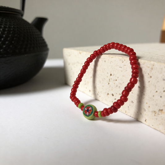 Red beaded bracelet with a colorful bead on a white surface with a black teapot in the background