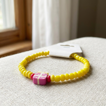 Yellow beaded bracelet with a pink and red square charm on a textured surface.