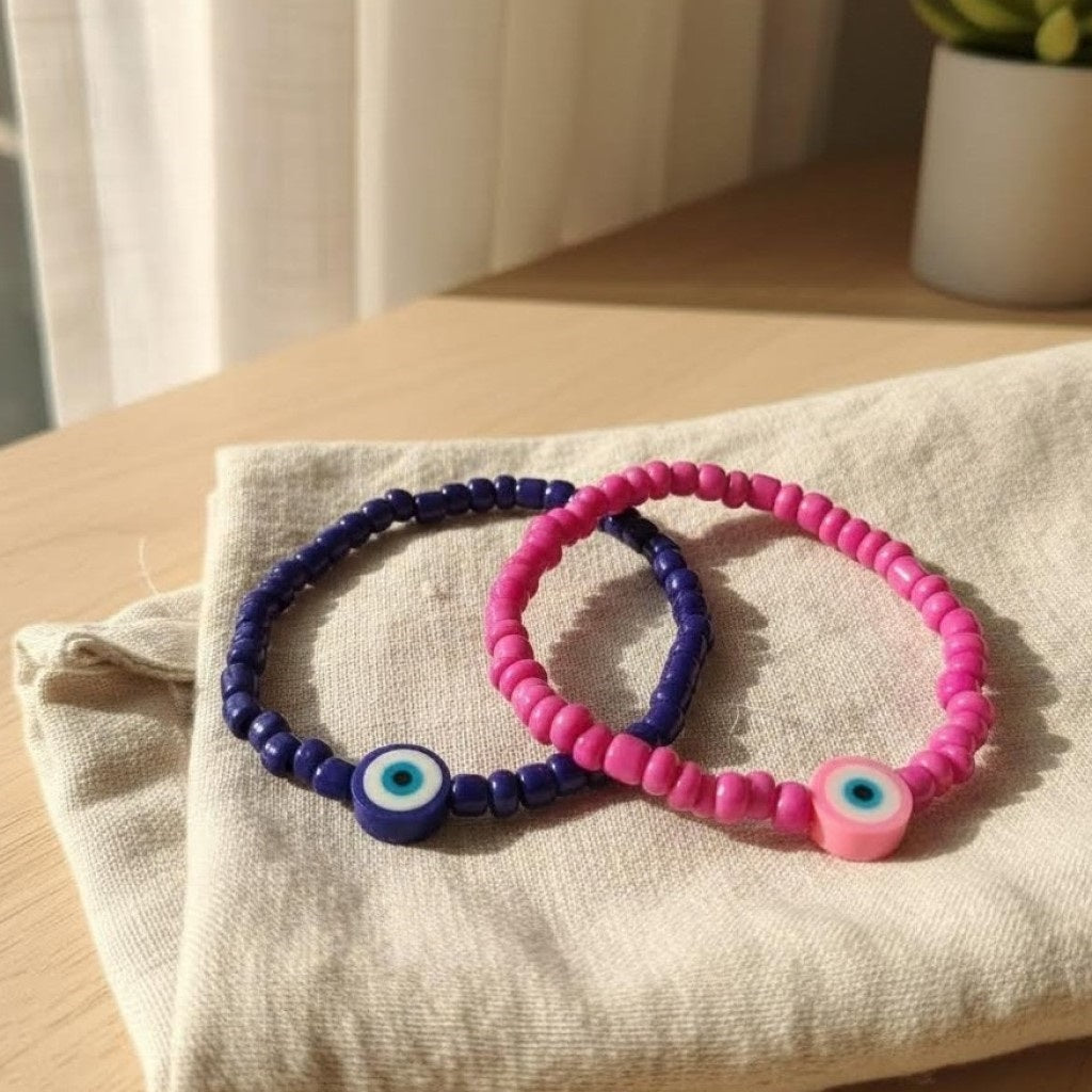 Two beaded bracelets, one pink and one blue, with eye-shaped charms on a light fabric surface.